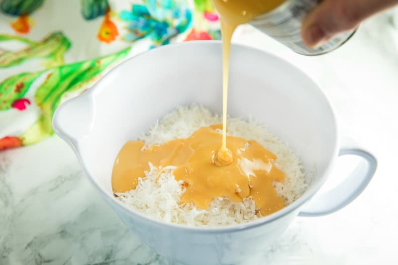 Adding condensed milk to coconut in a white mixing bowl