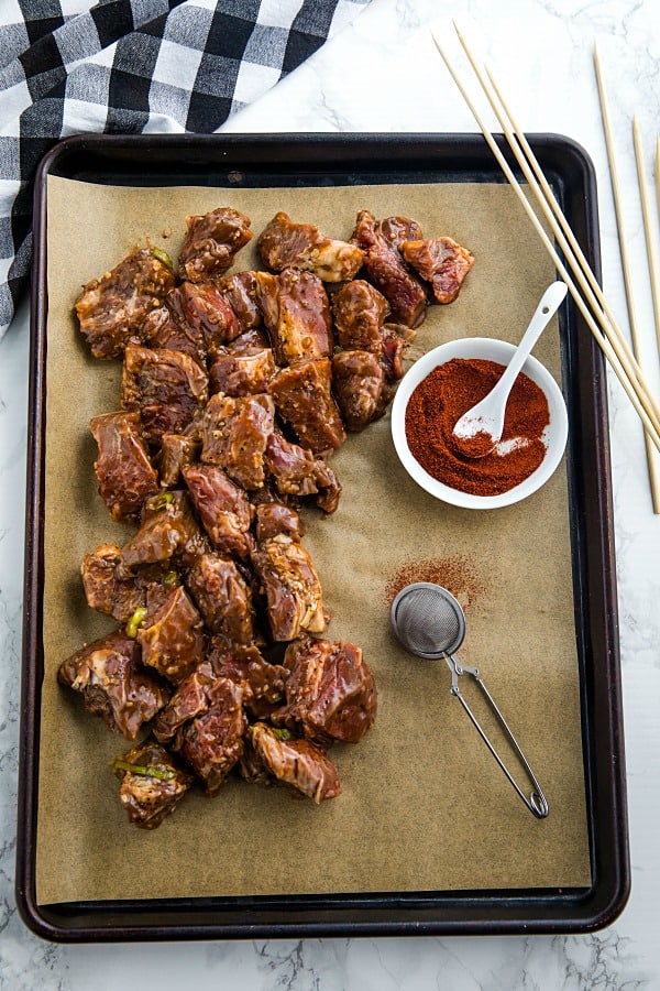 Marinated beef chunks on parchment lined tray with chili powder and smoked paprika mixed in small white bowl